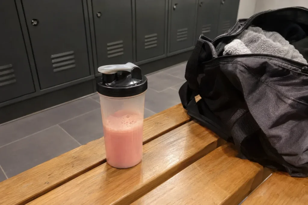 Shaker de whey fraise et sac de sport posés sur un banc de vestiaire